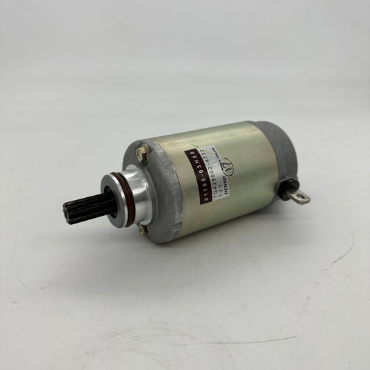A small cylindrical electric motor with a brushed silver metal body, a black splined shaft on one end, and a small metal terminal on the other, resting on a light gray surface.